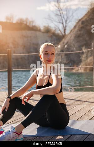 Una donna si prende una pausa dal suo allenamento, seduta su un tappetino vicino all'acqua sotto la luce del sole. Foto Stock