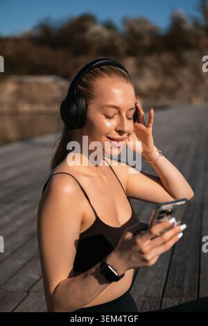 Una donna ama ascoltare musica con le cuffie mentre si allena all'aperto, in riva all'acqua e alla luce del sole. Foto Stock