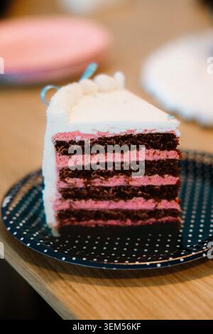 Primo piano di una fetta di torta al cioccolato a strati e alla crema di lamponi rosa con glassa bianca e nastro blu su un pallino di polka blu Foto Stock