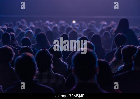 Grande pubblico seduto a guardare lo spettacolo sul palco in un luogo buio, l'illuminazione blu crea un'atmosfera soffusa. Foto Stock