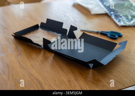 Confezione regalo in cartone nero opaco su un tavolo in legno alla luce del sole con forbici blu. Concetto: Imballaggio e-commerce Foto Stock