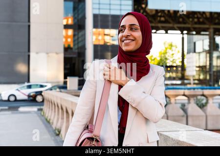 Donna d'affari del Medio Oriente araba che indossa un hijab passeggiando per la città - giovane donna musulmana adulta che cammina con una borsa in un ambiente moderno della città Foto Stock