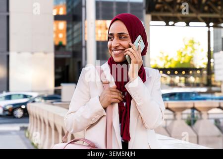 Donna d'affari del Medio Oriente araba che indossa un hijab passeggiando per la città: Donna d'affari sicura che sorride, parla con lo smartphone mentre cammina nell'area industriale Foto Stock
