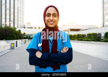 Medico sorridente del Medio Oriente con uno stetoscopio e scrub in piedi fuori da un edificio moderno, che rappresenta professionalità e dedizione in salute Foto Stock