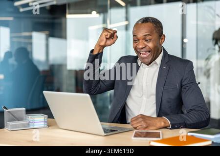 Uomo d'affari maturo che prova gioia e trionfo, alzando il pugno in un gesto di celebrazione mentre guarda lo schermo del suo notebook, ottenendo il successo professionale in un ambiente d'ufficio moderno Foto Stock