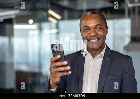 Uomo maturo che tiene in mano uno smartphone, guarda la fotocamera con un'espressione gioiosa, simboleggia la comunicazione di successo, la tecnologia moderna e la connettività aziendale in un ambiente professionale Foto Stock
