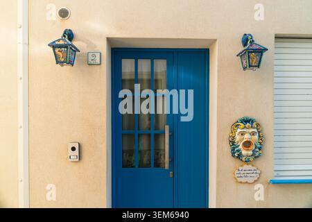 Vista di una porta in legno blu vibrante con pannelli di vetro, lanterne decorative e una maschera in ceramica su una parete beige a Marsaskala, regione meridionale, Malta. Foto Stock