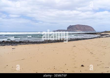Una tranquilla spiaggia a la Graciosa, Lanzarote, caratterizzata da una spiaggia sabbiosa, onde oceaniche e un'isola lontana. Foto Stock