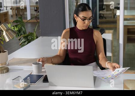 Donna asiatica che indossa la parte superiore Bordeaux, lavora su un laptop alla scrivania, esamina le carte e tiene in mano la tazza Foto Stock