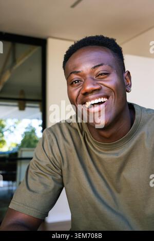 Uomo africano sorridente a metà degli anni '20 che indossa T-shirt Olive e orecchini a cerchio sul patio vicino alla porta di vetro Foto Stock