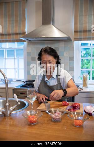 Donna cinese anziana che taglia verdure sull'isola della cucina con grembiule grigio e coltello da cuoco a bordo Foto Stock