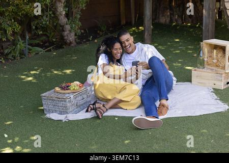 Coppia afroamericana che ride, che si piega insieme mentre guarda lo smartphone sulla coperta da picnic Foto Stock