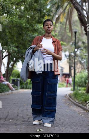 Donna africana che cammina sul sentiero lastricato del parco tenendo in mano la felpa grigia, indossando occhiali e scarpe da ginnastica Foto Stock