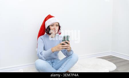 Woman in Babbo Natale si siede su un tappeto accogliente, interagendo con gli amici tramite smartphone durante i festeggiamenti di Capodanno, irradiando vivaci emozioni natalizie Foto Stock