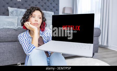 Ottawa ON, Canada - 6 ottobre 2025: Donna con i capelli ricci seduta sul pavimento, tenendo in mano un laptop che mostra Netflix cancellata, circondata da un accogliente salotto Foto Stock