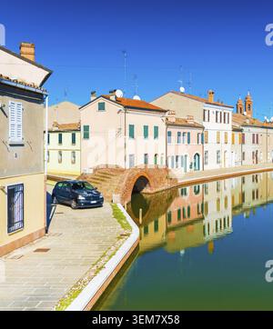 Una strada della piccola città italiana di Comacchio. Case colorate riflesse nell'acqua. Ponte in pietra e canale d'acqua in stile veneziano Foto Stock