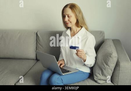 Donna felice sorridente che tiene in mano una carta di credito di plastica, guarda il computer portatile, acquista online o ordina prodotti tramite computer seduto sul divano in l Foto Stock