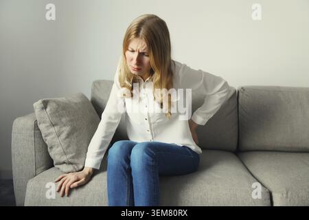 Una giovane donna che soffre di mal di schiena, tocca la schiena mentre si siede sul divano nel soggiorno di casa Foto Stock