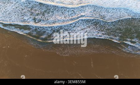 La vista dall'alto cattura le onde azzurre schiumose che rotolano sulla sabbia bagnata, formando linee naturali, riflessi e texture lungo la costa Foto Stock