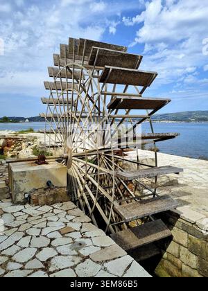 Vista di un'antica ruota ad acqua intemprata su uno sfondo di acqua e un cielo parzialmente nuvoloso. La ruota è costituita da pale in legno supportate da un metallo Foto Stock