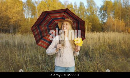 Donna felice nel parco autunnale con ombrello che tiene in mano foglie d'acero, gode del clima caldo, guarda lontano, alberi con foglie gialle, sfondo naturale della foresta Foto Stock