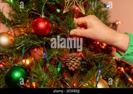 Primo piano di una mano che appende un ornamento a cono di pino dorato su un albero di Natale decorato con luci festive colorate Foto Stock
