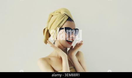 Ritratto di una donna sorridente di stile che si asciuga i capelli bagnati con un asciugamano avvolto sulla testa e si diverte con gli occhiali da sole alla moda su sfondo grigio Foto Stock
