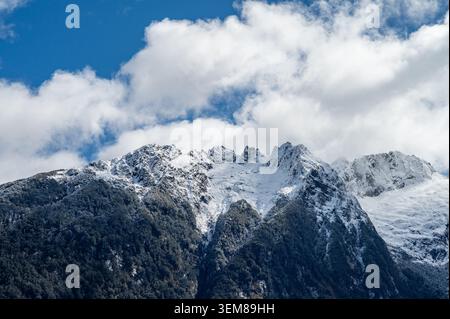 Vista ravvicinata di una spettacolare e aspra vetta della montagna Remarkables coperta di neve e ghiaccio sopra una foresta verde scuro sotto un cielo blu nuvoloso. Foto Stock