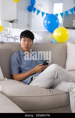 Uomo adulto asiatico seduto su un divano beige a casa con uno smartphone che indossa una camicia blu con palloncini Foto Stock