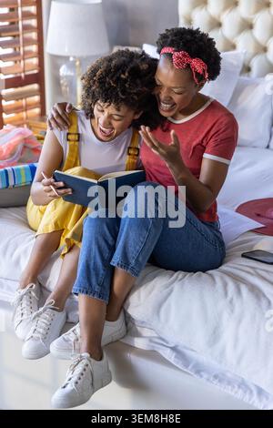 Due amiche sedute a letto in camera da letto, leggono un libro aperto e ridono con lo smartphone nelle vicinanze Foto Stock