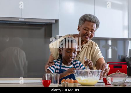 Donna africana anziana che guida la ragazza africana che mescola la pastella nella ciotola di miscelazione sull'isola della cucina Foto Stock