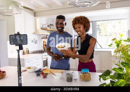 Coppia afroamericana che mostra la ciotola di granola al telefono su un treppiede in una cucina luminosa con la parte superiore del raccolto Foto Stock