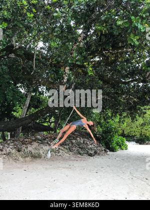 Giovane donna felice con un costume da bagno che oscilla su una corda appesa a un lussureggiante albero tropicale sopra una spiaggia di sabbia bianca incontaminata. Momenti di vacanza divertenti. Foto Stock