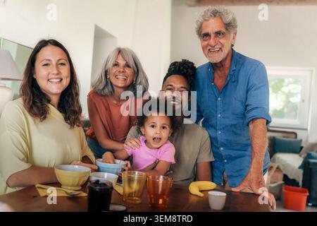 Famiglia plurirazziale felice con nonni e bimbi piccoli che gusteranno la colazione insieme a casa. Autentico stile di vita di tre generazioni con un sorriso Foto Stock