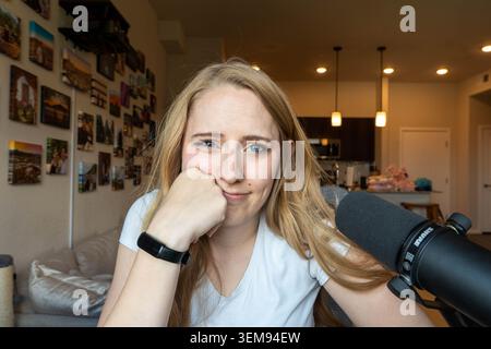 Una giovane donna con i capelli biondi sorride alla telecamera, appoggiando il mento sulla mano in un accogliente studio domestico. Un microfono di grandi dimensioni si trova su una scrivania di fronte Foto Stock