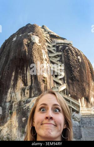 Una donna bionda con un sorriso sorpreso si erge di fronte a una formazione rocciosa torreggiante, con una scalinata di legno che sale sulla scogliera e un cielo blu brillante Foto Stock