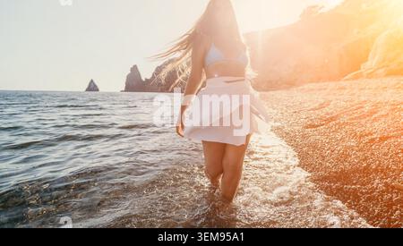 Donna in spiaggia che cammina. Elegante giovane donna in gonna bianca che passeggiava lungo la riva di ciottoli al tramonto durante le vacanze estive Foto Stock