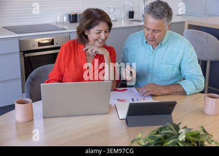 Camicetta rossa donna anziana e camicia blu maschio matura che rivede laptop e grafici al tavolo della cucina Foto Stock