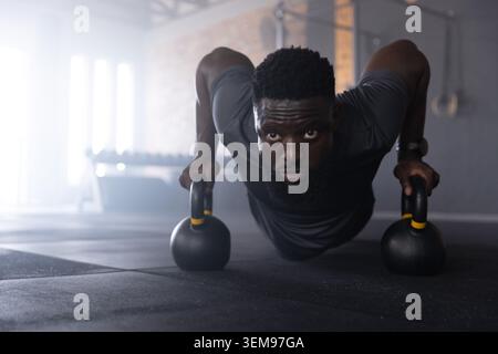 Uomo afroamericano che fa push-up kettlebell sul pavimento della palestra indossando camicia atletica scura e orologio da polso Foto Stock