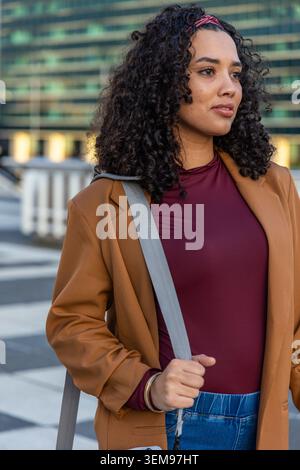 Donna in piedi sulla terrazza sul tetto con soprabito in cammello, top marrone, pantaloni in denim e borsa a spalla Foto Stock