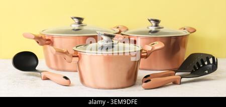 Moderno set di pentole in rame con pentole, pentole, coperchi in vetro e utensili da cucina su un tavolo bianco su sfondo giallo Foto Stock