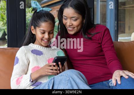 Madre e bambino asiatici seduti vicino al soggiorno, condividono il telefono nero sul divano in pelle marrone Foto Stock