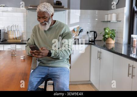 Un uomo afroamericano anziano seduto in cucina a controllare lo smartphone indossando il top di salvia e un orologio nero Foto Stock