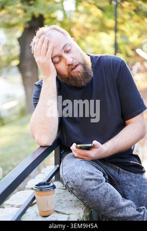 Un uomo caucasico stanco è sbalzato sulla panchina tenendo lo smartphone e caffè da asporto, barba e camicia nera informale, alberi autunnali illuminati dal sole sullo sfondo, un lavoratore remoto in attesa di un messaggio nel tardo pomeriggio, schietto Foto Stock