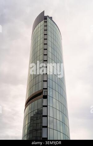 Alto edificio di vetro che si piega verso l'alto in un cielo nuvoloso, riflettendo l'architettura circostante Foto Stock