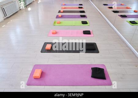 Lo studio di yoga vuoto presenta file di tappeti vivaci, asciugamani piegati neri e blocchi di yoga corallo su pavimento di legno chiaro sotto la luce naturale, che si riflettono Foto Stock