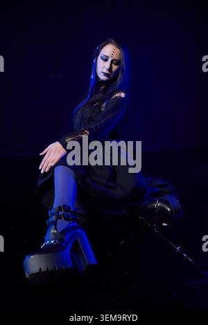 Giovane donna gotica dai lunghi capelli blu tinti e un trucco spettacolare si siede sul pavimento con un abito in pizzo nero e grossi stivali con plateau sotto una moody ligh blu Foto Stock