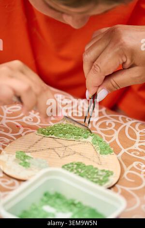 Vista dettagliata della persona che applica con cura piccole piastrelle a mosaico verde su un asse di legno utilizzando pinzette su tovaglie a motivi sotto un'illuminazione calda Foto Stock