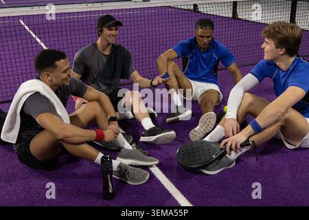Diversi giocatori maschili in abbigliamento sportivo seduti su un campo viola vicino alla rete, tenendo le pale e la palla. Racchetta, cameratismo, attività ricreative, palestra, candid, un lavoro di squadra vivace Foto Stock