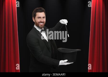 Mago che esegue trick con bacchetta e cappello sul palco contro tende rosse Foto Stock